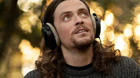 Chris Maddaloni A long-brown haired man looks to the sky. He is surrounded by green shrubbery. He wears black headphones, a black t-shirt and a blue jumper.