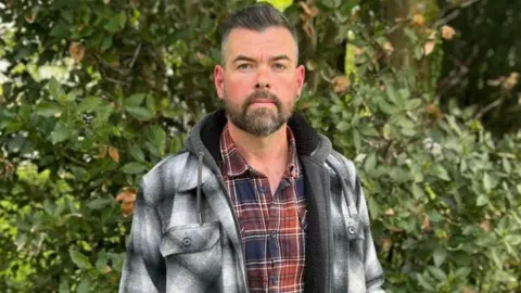 A man wearing a black and white checked jacket and red and blue checked shirt stands in front of shrubbery. He has a short beard, dark in colourand short dark hair. He is looking directly at the photographer.