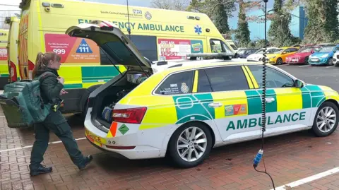 Chrissy Ames has a very large, green backpack over one shoulder. She is about to load it into the open boot of her ambulance estate car. The car is parked beside a row of ambulances at the Kidlington ambulance station. Black and curly power cables are hanging from above.