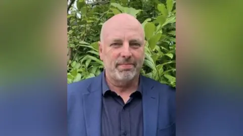 A man in a blue shirt and jacket standing outside in front of some greenery