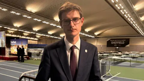 A man stands in a leisure centre where tables are in the background for the vote count. He has a navy suit, burgundy tie, brown short hair and black square glasses. He has a neutral expression but looks sad.