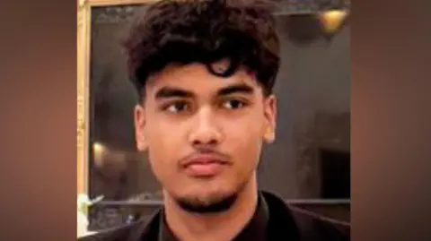 Absi Ahmed looking to the right of the camera. He has dark, curly hair which is short on the sides and long on the top. The photo is cropped close, with only his top and shoulders visible. He is wearing a black suit jacket, black shirt and black tie.