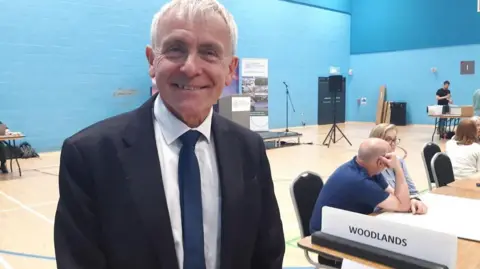 LDRS/Anttoni James Numminen A smiling grey haired man in a blue jacket and tie and white shirt stares at the camera