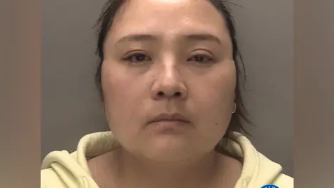 A mugshot of a woman who has dark hair and brown eyes, wearing a yellow hoodie