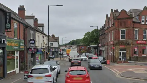 A road with shops on both sides and lots of cars travelling in both directions. Cars parked on both sides of the road and a bus stop on the left hand side.