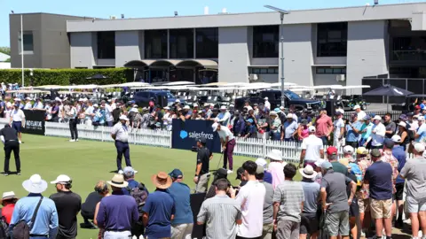 Sarah Reed/Getty Images An image featuring a putting green in Adelaide, Australia, and including a man in a white cap, a white top and purple trousers holding a golf club. People are watching behind a white fence and in front of a building in the top half of the image and others are watching at the bottom of the photo.