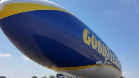 The blue-and-yellow Goodyear blimp moored on an airfield as people gather near the passenger cabin before or after a flight.