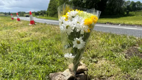 A bouquet of yellow and white flowers wrapped in plastic wrap are placed vertically held down with stones at the roadside. In the distance are some orange cones placed by the side of the road.