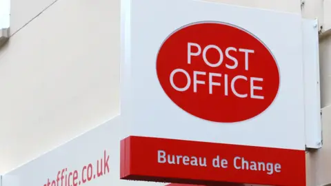 A white and red Post Office sign hangs from the side of a building. The words "Post Office" in white writing sit within a red circle. Below are the words "Bureau de Change", in similar white writing enclosed in a red rectangle. 