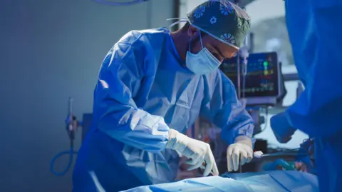 A stock image of a surgeon in blue scrubs leaning over a patient holding tools.
