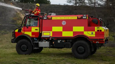 An off-road fire engine with large tires and a red and yellow livery. Surrey Fire and Rescue Service is written along the top of the vehicle. A man is spraying water from a hose above the cab.