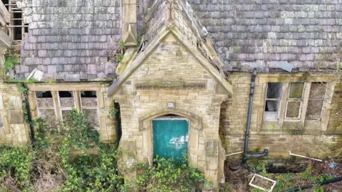 A rundown school building wih a green door and lots of overgrowth around the front. There is visible damage to the roof. 