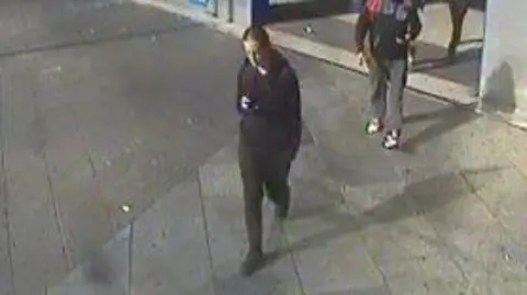 Met Police A CCTV still of Alfie Coleman wearing a black hoodie and trousers while on his phone. He is walking with automatic doors open and another man behind him.