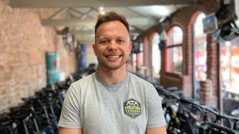 Patrick Kozlowski is a man with brown hair. He is standing in a bike showroom with two rows of bikes behind him. He is wearing a grey t-shirt that has the logo of his bike shop on it.