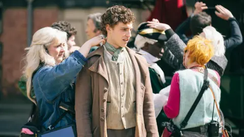 Dan Ollerhead Sánchez outside Wentworth Woodhouse. He is wearing period‑style clothing while several crew members work closely around him. One crew member on the left is adjusting the collar or scarf at his neck, holding it carefully with both hands. Another crew member on the right, wearing bright clothing and carrying equipment, appears to be preparing or checking costume details. Additional people are visible in the background, some wearing hats and period costumes, while others appear to be part of the production crew.
