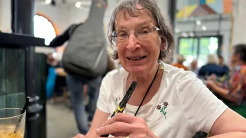 Mairead Kirwin is an older lady, with short grey hair. She is smiling, wearing glasses, a white t-shirt and holding a penny whistle, sitting in a busy cafe. 