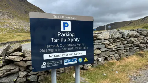 A sign in the Kirkstone Pass car park notifying users they are on private land. It reads: "Parking tariffs apply, terms and conditions apply, see signs in car park for details, no cash payment option". It is positioned in front of a dry stone wall. Hills can be seen in the background.