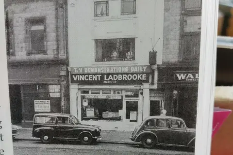Johnny Haynes Two vintage cars parked in front of a shop with signage saying TV demonstrations daily, Vincent Ladbrooke