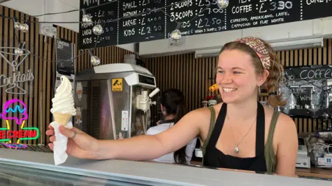 A woman smiles as she passes over an ice cream in a cone to a customer from a market stall in Norwich. She wears an apron and a headband. 