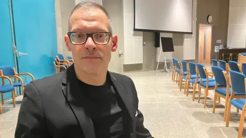 BBC/Amy Holmes Independent Central Bedfordshire councillor Adam Zerny, who has glasses and is wearing a black suit jacket and a black t-shirt. He has short grey hair. He is sitting down. There are rows of blue and brown chairs in the background.