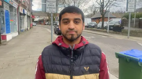 Man with dark hair wears a black and yellow body-warmer over a red jacket while standing in a street with shop fronts on the left and a road on the right. A green wheelie bin is in the bottom-right corner.