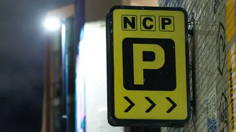 The logo of an NCP car park, which is rectangular, yellow and black, attached to a grey brick wall that has graffiti on it. It is dark and the background is blurred.