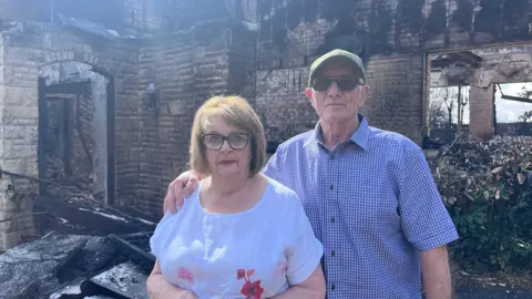 A woman with brown hair, glasses and a white patterned T-shirt next to a man with a green cap, blue shirt and sunglasses with one arm over her shoulder and the remains of their fire-damaged home behind them