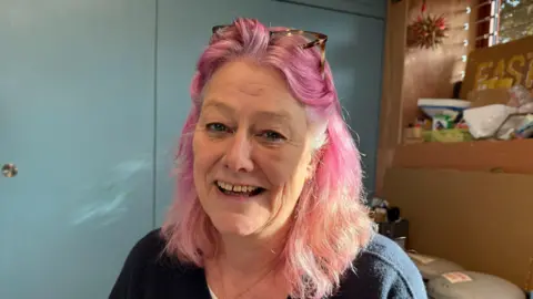 Woman looking at the camera. She has pink hair and glasses on her head. She is smiling at the camera.