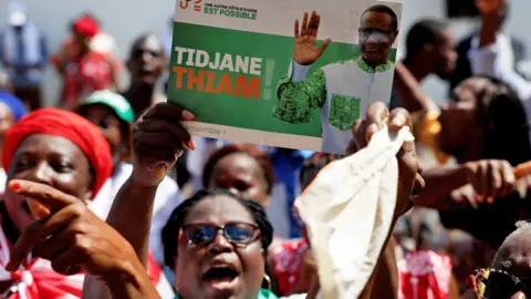 Female PDCI supporters protest against a court judgement to remove their leader Tidjane Thiam from the electoral list in Abidjan, Ivory Coast. One holds up a campaign poster of Thiam waving -  24 April 2025