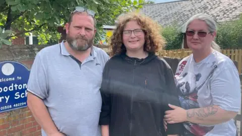 Andrew Turner/BBC John, Jesse and Donna stand together near the school sign, that reads: Welcome to Woodton Primary School, giving details of the phone number. They are all wearing casual clothes, standing near a brick wall and fence, with trees and hedges nearby.