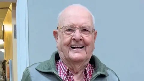 Jack Waterfield smiling at the camera, with a glass of fizz in his hand. He has short white hair and glasses, and is wearing a green zip-up top over a white-and-red checked shirt. He is in a room with some people in military uniform around him.