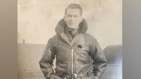 Shaun Whitmore/BBC A black and white 1940s or 1950s photo of George Kelly. He is in uniform, wearing a leather flying jacket done up with a zip and with its fur collar raised around his neck. His hands are in his pockets. Behind him can be seen corrugated Nissan huts.