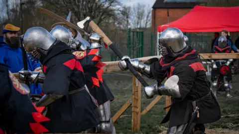 David Polier Two people dressed in medieval outfits engaged in combat. One is striking the other with an axe