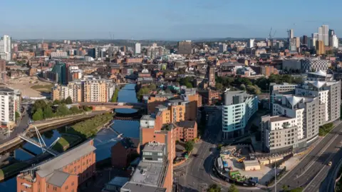 An aerial photograph of Leeds showing an urban skyline and a canal running through the middle. There is a mix of modern high-rise buildings and older brick structures.