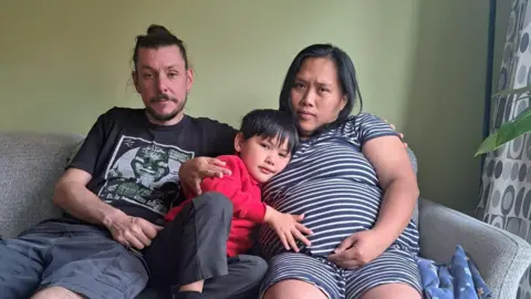 Neil and Ferni Evans, with their son Johny. The family are sitting on a sofa, looking worried.