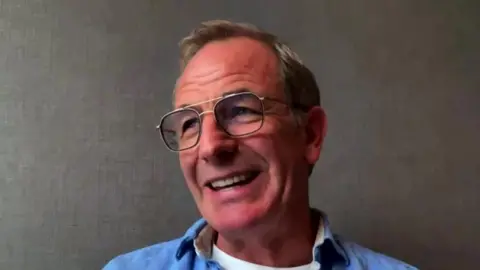 Robson Green during a video call. He is wearing a blue shirt and glasses.