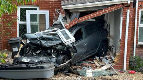 The front of a smashed up grey car which can be seen with its rear crashed into the front of a house. The door of the house is destroyed and the porch of the house is hanging down. There is a lot of debris on the driveway of the house. 