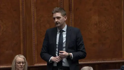 NI Assembly Phillip Brett speaking in the assembly. He has short brown hair, wearing a navy blazer, light blue shirt, navy tie and a watch. Wood panelling is behind him and another woman with blonde hair and glasses is seated next to him.