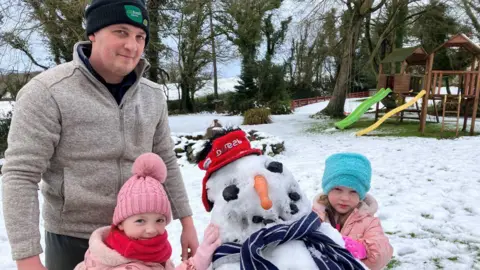 Matthew Craig aparece en la foto con sus dos hijos y un muñeco de nieve que construyeron. Todos llevan gorros y suéteres, y las niñas llevan abrigos rosas. Detrás de ellos hay una zona de juegos.