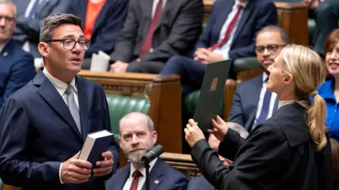Andy Burnham, in a suit and tie, holds a Bible and reads from a card held up by a female Commons official in the House of Commons chamber