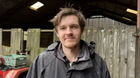 Chris Powell stands in front of a wooden fence. He is wearing a dark jacket specked with rain. He has light brown hair and a short beard.