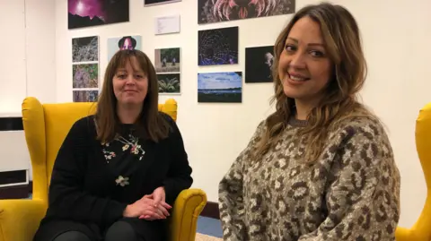 Jon Wright/BBC Michelle and Charlotte sit on two yellow arm chairs in front of photography exhibition inside a library