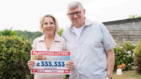 Postcode Lottery Rebecca Andrews and her husband David Docherty hold a large check with £333,333 on it. They are standing on a grassed lawn surrounded by shrubs and bushes.