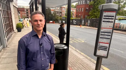 A man in a blue long sleeved-top standing by a bus stop on Golders Green Road.