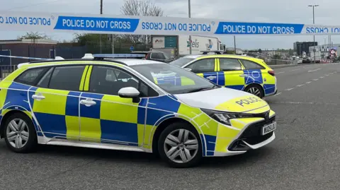 Two police cars with blue and white livery are parked close to a line of blue and white police tape on a road