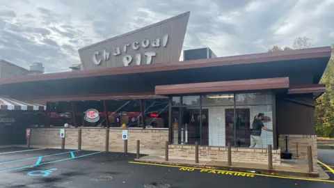 The Charcoal Pit diner is brown with windows along the brown, a sign pokes out from the top of the building with the restaurants name on it in large white bubble writing. The image is taken from the car park where a disability space is seen and a yellow 'no parking' area is highlighted.