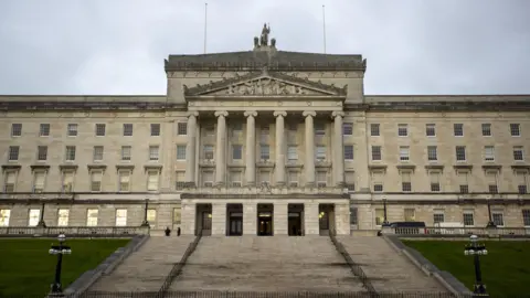 PA Media Parliament Buildings at Stormont Estate. It is a Greek classical style building with steps leading up to it and lawns on either side.