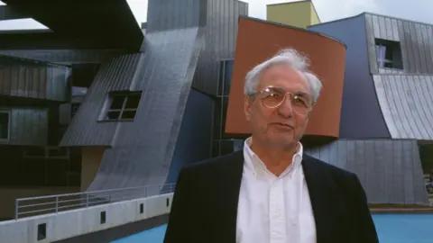 A man in a white shirt and black jacket and wearing glasses stands in front of a building. He has white hair