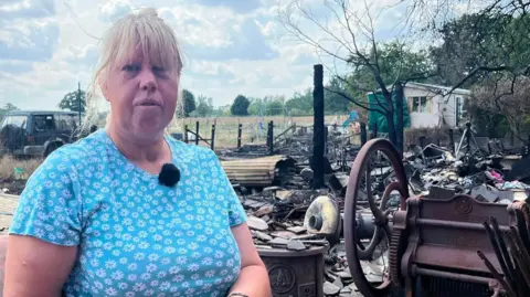 Hannah Gray/BBC A blonde woman in a blue shirt standing in front of ash and rubble.