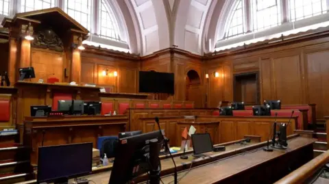 The interior of a courtroom. There's various computer screens, microphones red backed chairs and brown wooden benches. The court room is empty. 